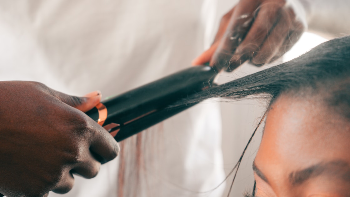 Hands straightening dark hair with a flat iron.
